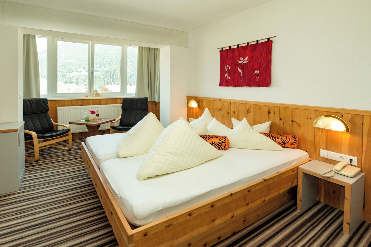 A hotel room with a large bed, two chairs, a small table, and a window with curtains.
