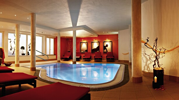 A swimming pool with red lounge chairs, a reception area with a mirror, and a decorative plant. - Hotel Alpina Resort (Wenns)
