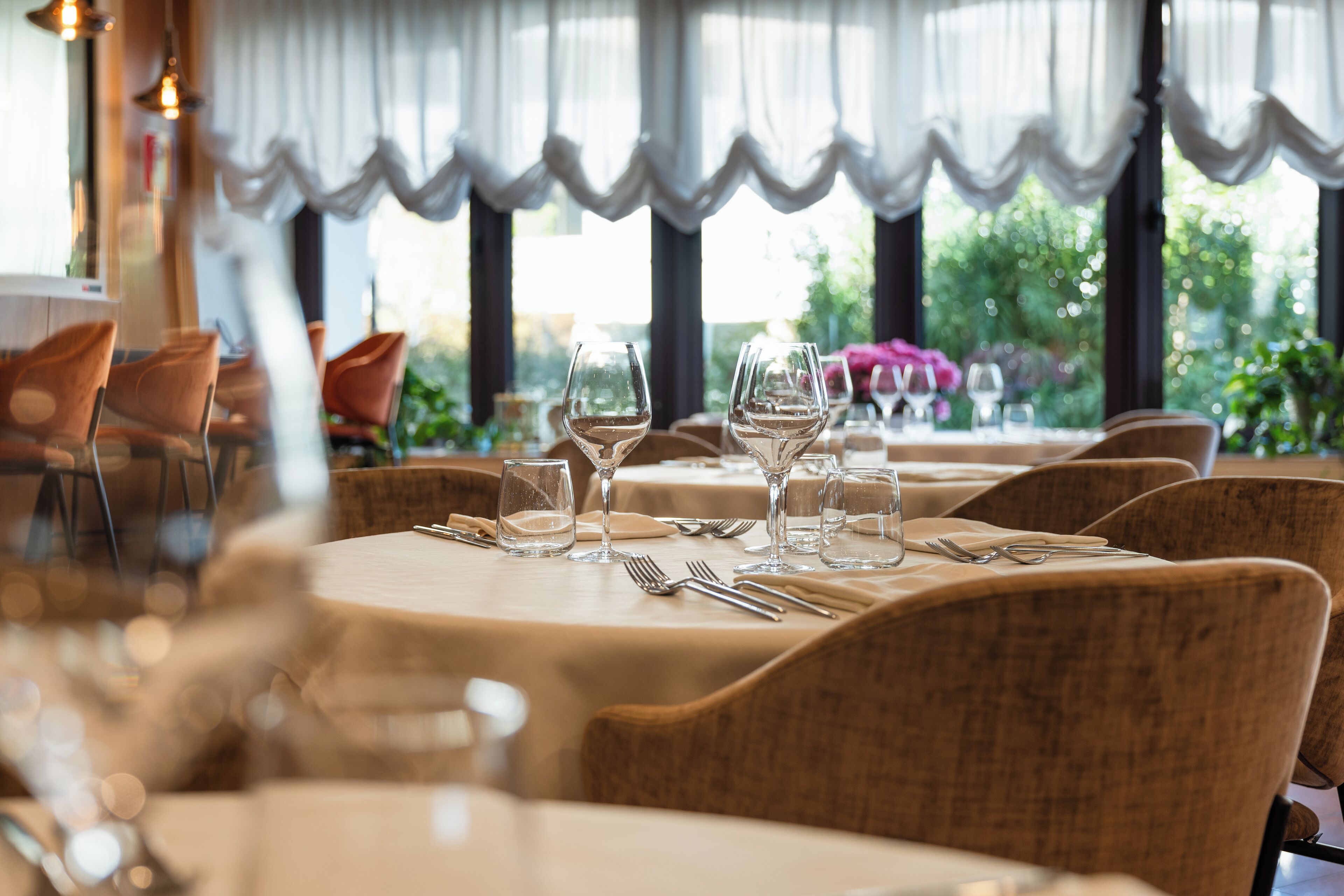 A restaurant with round tables, chairs, and empty wine glasses, with a view of greenery outside.