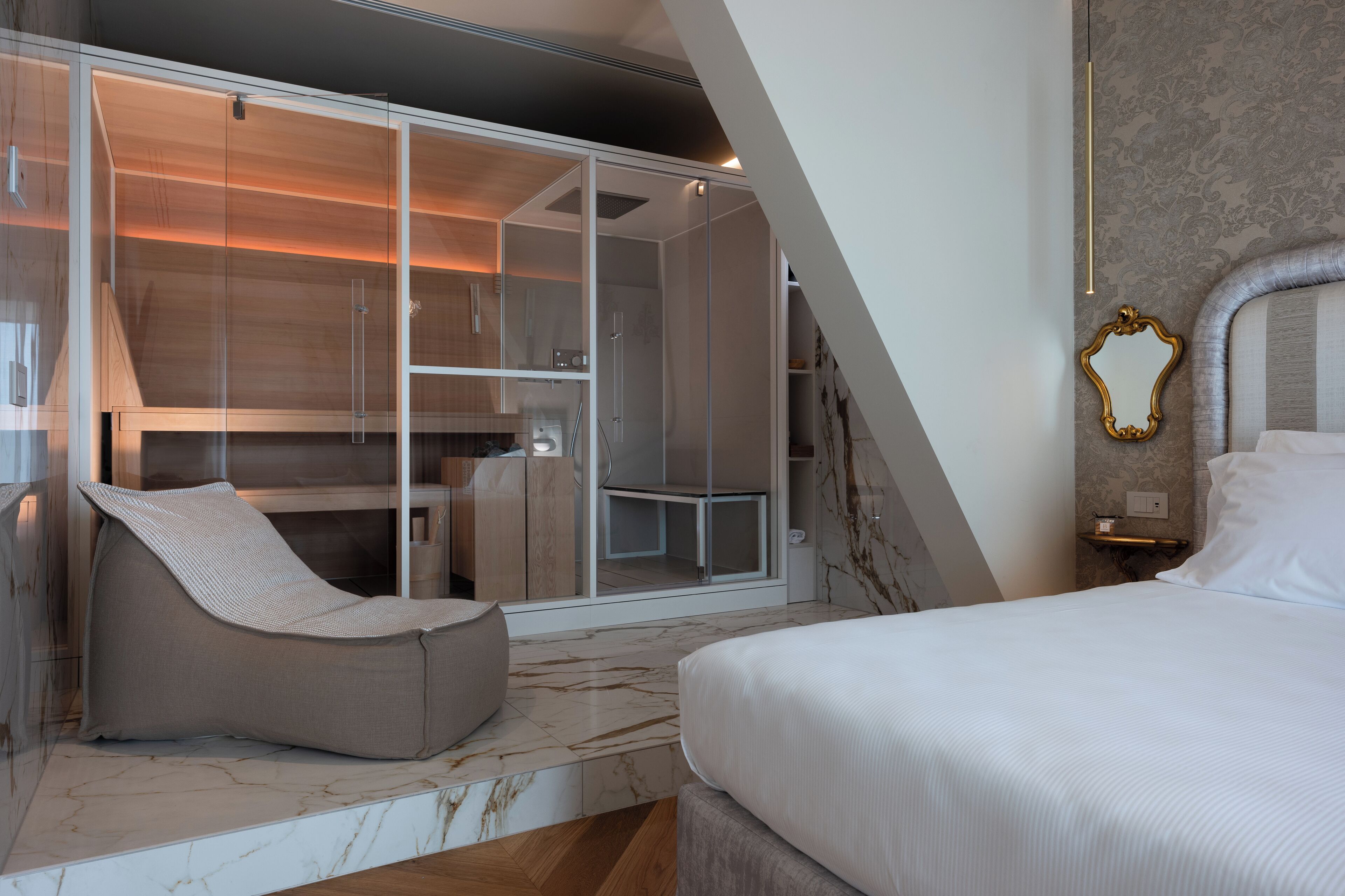 Un dormitorio moderno con una cama grande, un vestidor con puertas de vidrio, una zona de estar y un suelo de mármol.
