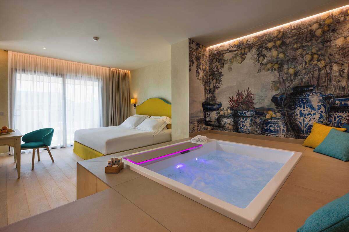 A hotel room with a bed, a massage table, a desk, and a large window with curtains.