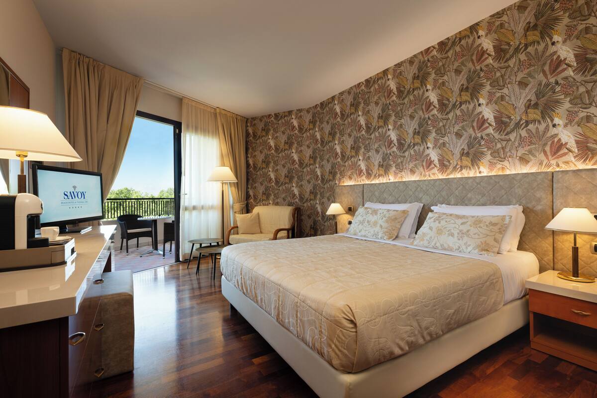 A hotel room with a large bed, a desk with a computer, a TV, and a balcony with curtains.