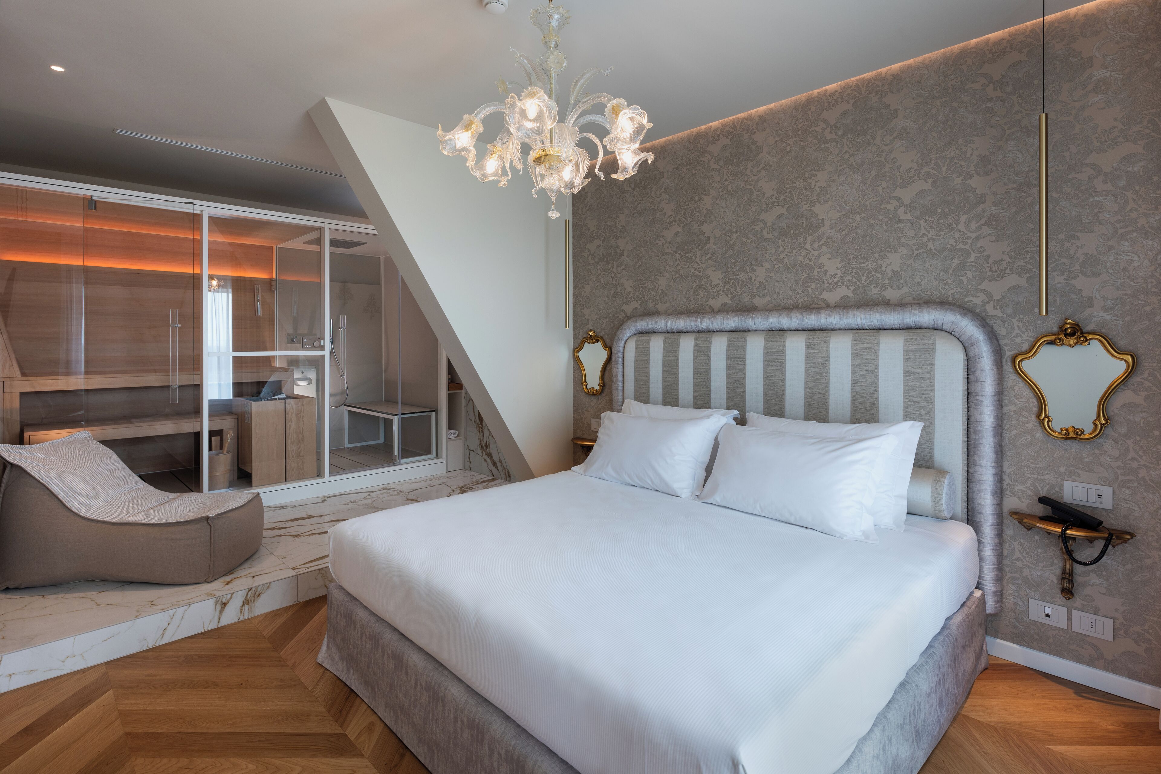 A modern bedroom with a large bed, a chaise lounge, a chandelier, and a view of a balcony.