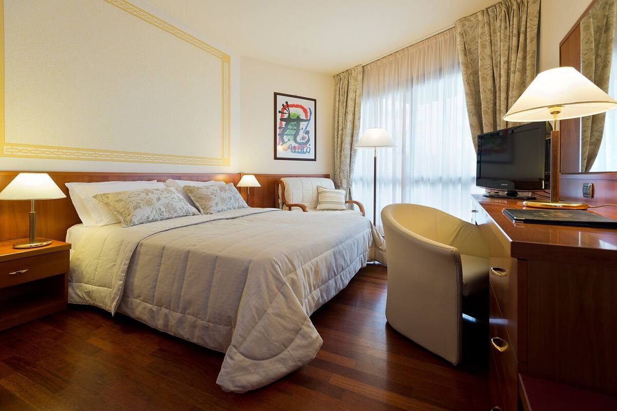 A hotel room with a bed, two bedside lamps, a desk with a television, and a chair.