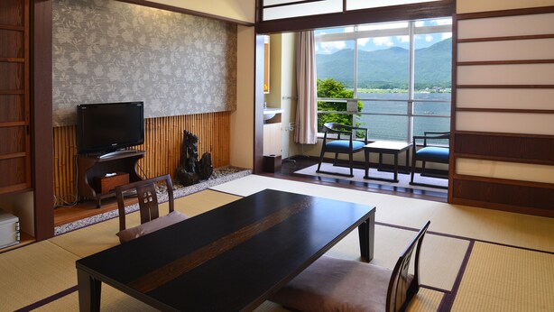 Une pièce de style japonais traditionnel, avec un sol recouvert de tatamis, une table basse, des chaises, un téléviseur et une vue sur un lac bordé de montagnes.