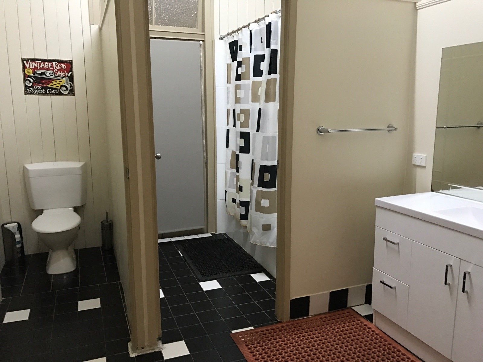 A bathroom with a toilet, a shower with a patterned curtain, a sink with a mirror, and a sign on the wall.
