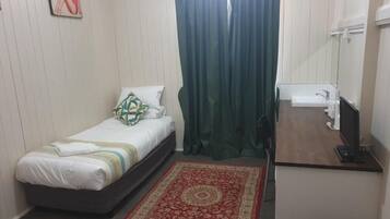 Un dormitorio pequeño y ordenado con cama, escritorio con computadora, una silla y un ventanal con cortinas.