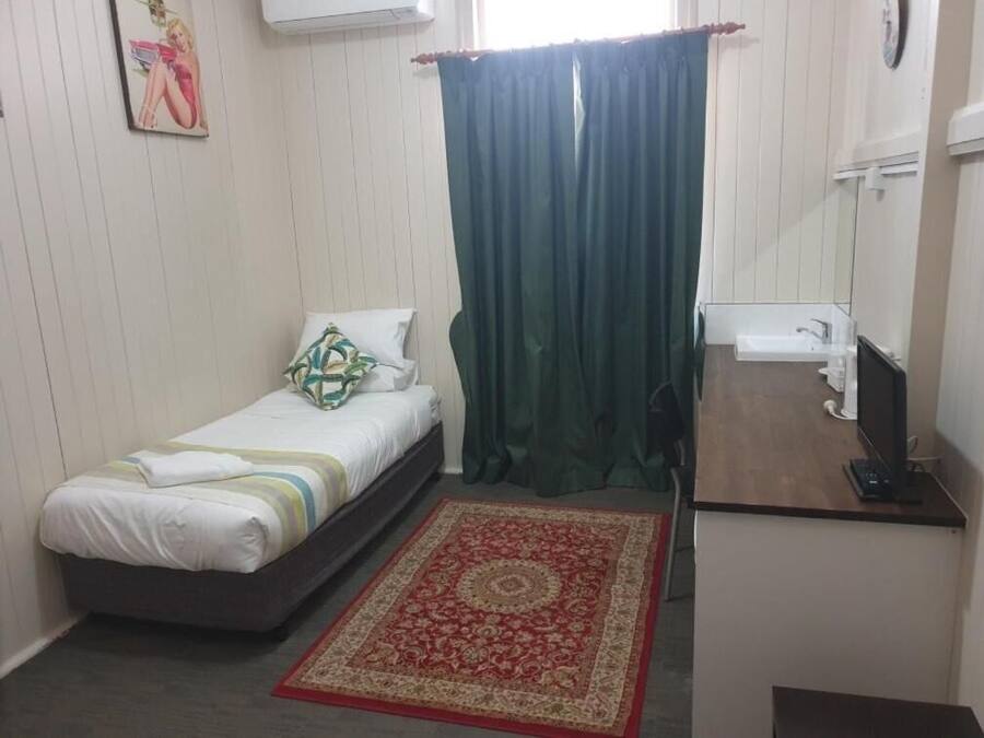 Un dormitorio pequeño y ordenado con cama, escritorio con computadora, una silla y un ventanal con cortinas.
