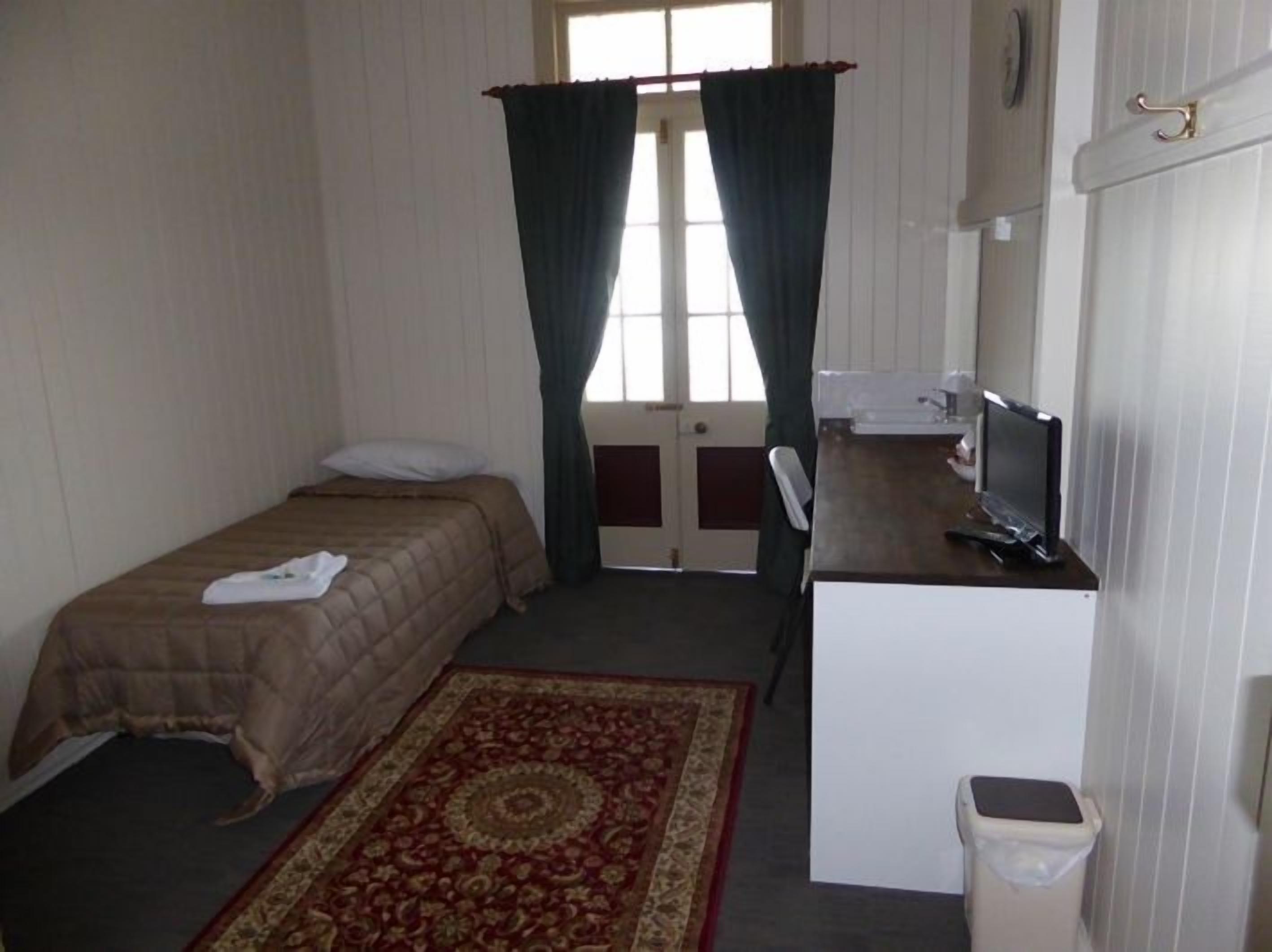 A small, simple room with a bed, a desk with a computer, a small trash bin, and a window with curtains.