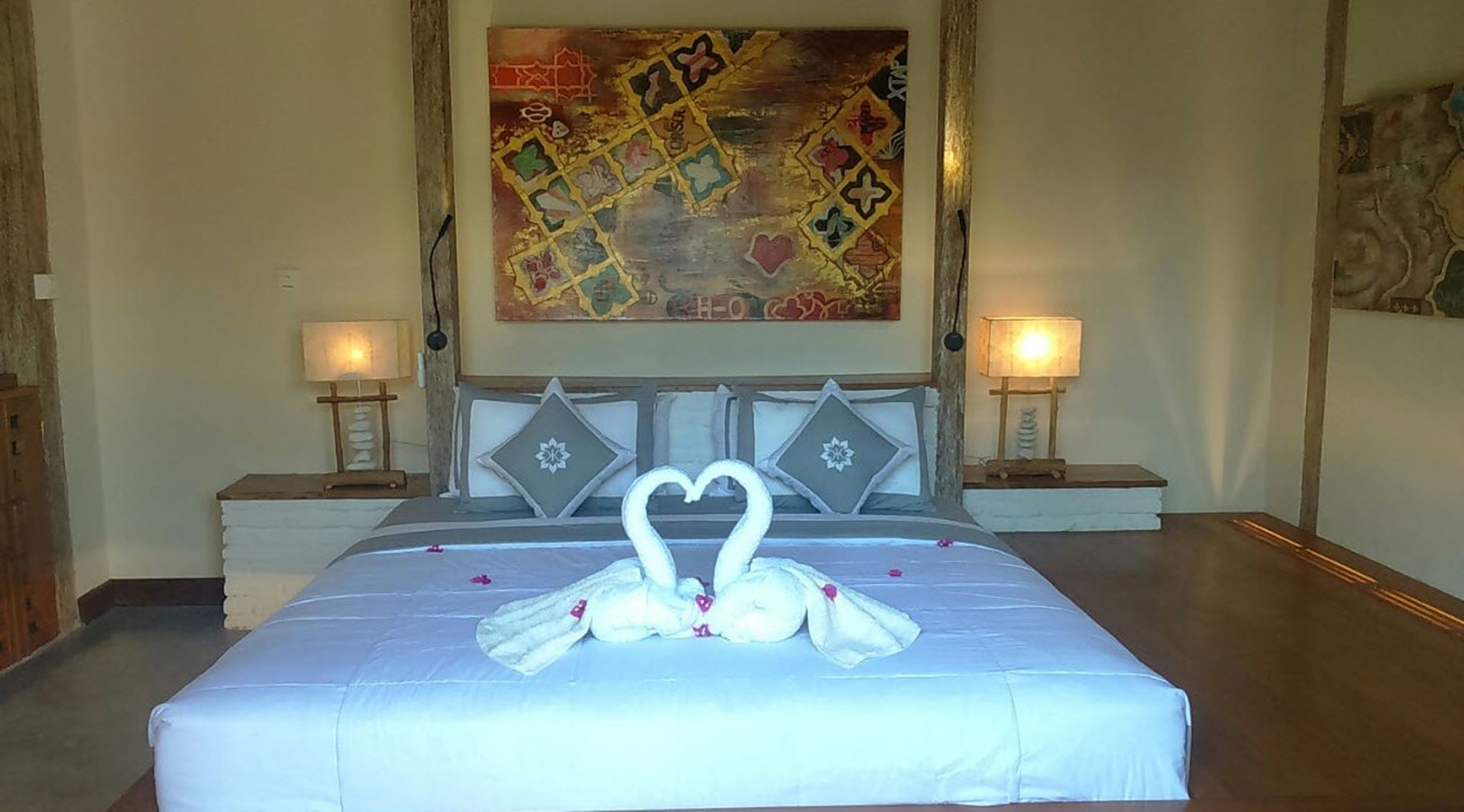 A hotel room with a large bed, two bedside lamps, a decorative wall piece, and a heart-shaped decoration on the bed.