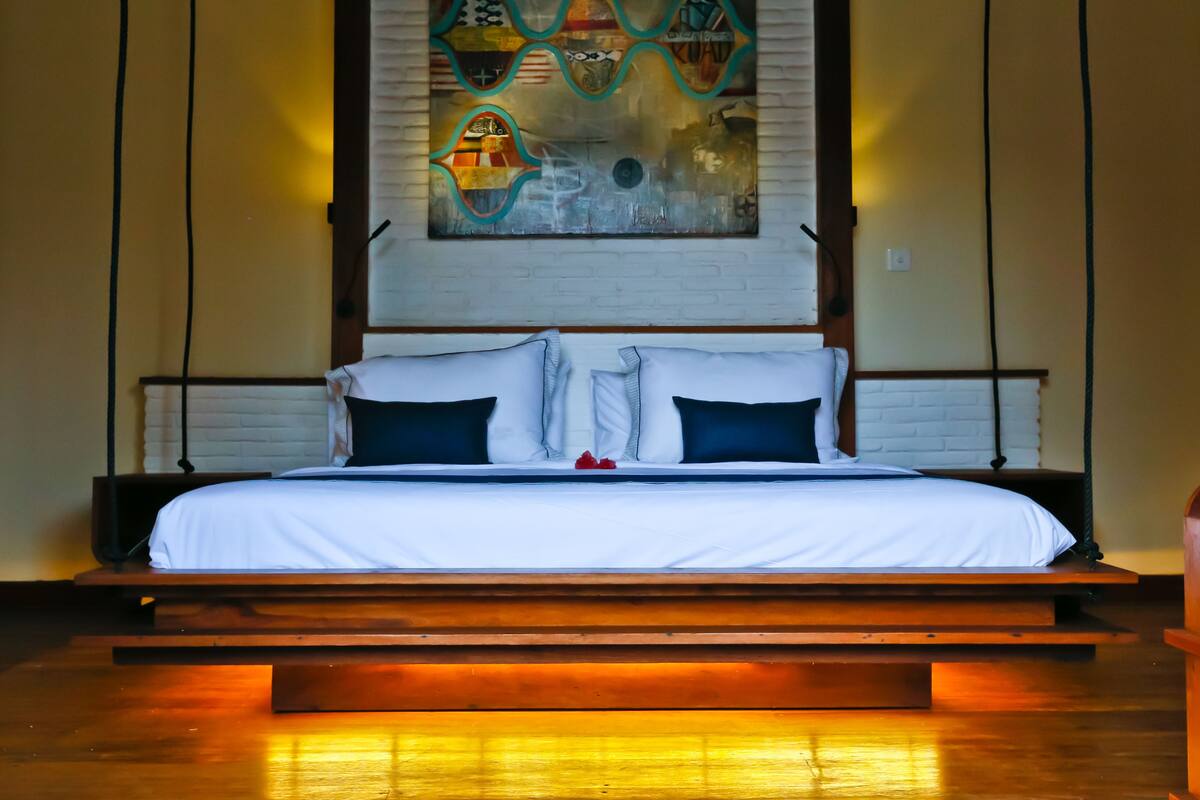 Een bed met een witte matras en donkere kussens, een houten hoofdbord en een aan de muur bevestigd abstract schilderij.