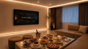 Une chambre d’hôtel moderne avec une table à manger dressée pour un repas, un canapé, un téléviseur diffusant des images de la marque de l’hôtel et une grande fenêtre avec des rideaux.