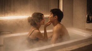 A couple in a bathtub with steam, a window with blinds, and a marble wall.