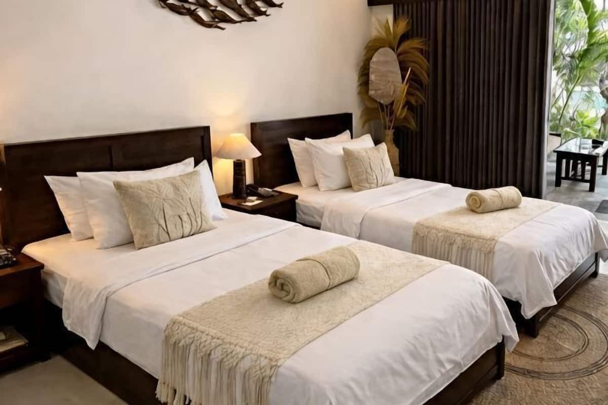 Dos camas individuales con ropa blanca y mantas color beige, un cabecero de madera, una mesita de noche con lámpara y una planta decorativa grande.