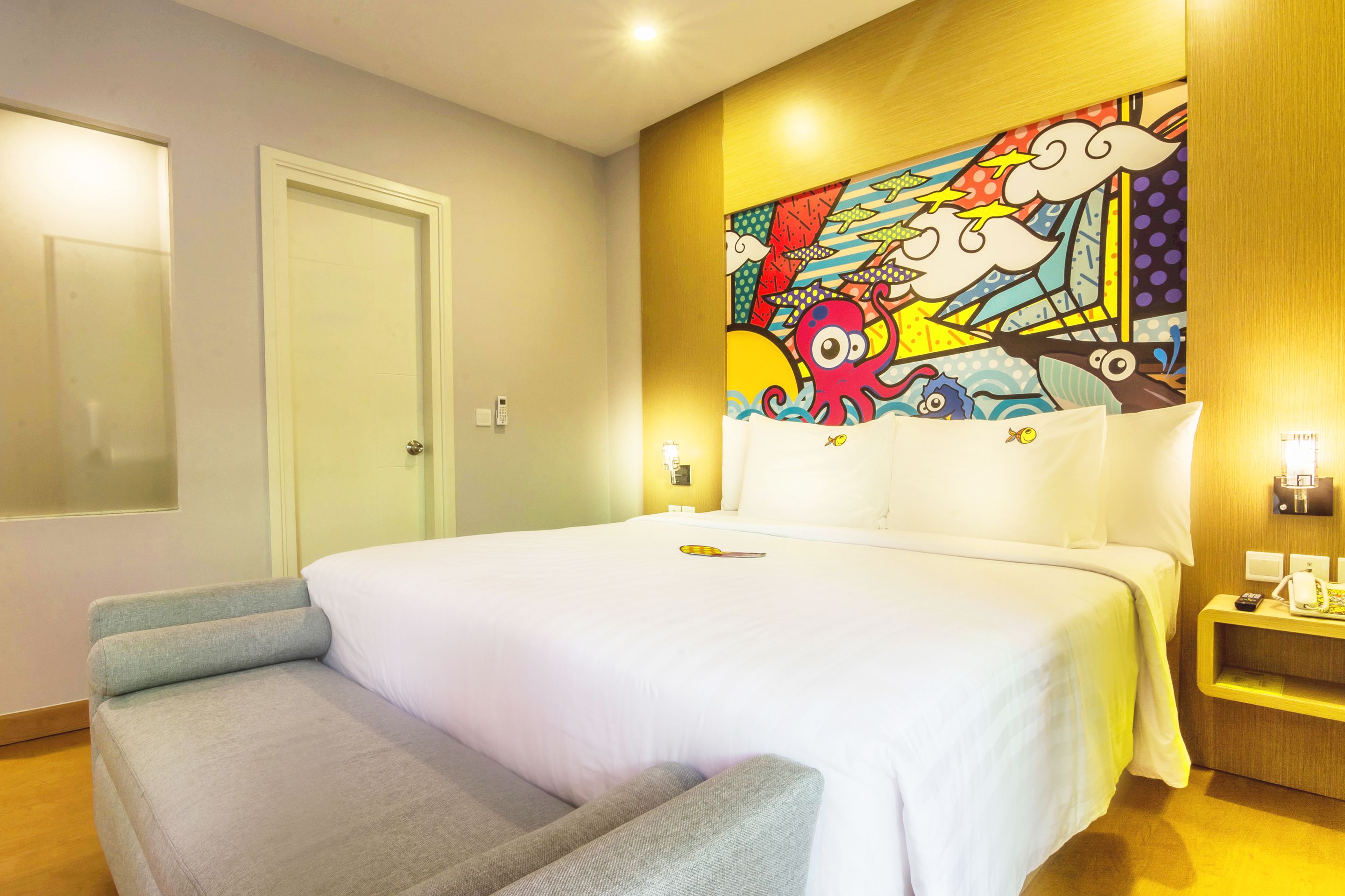 A hotel room with a bed, a chair, a colorful mural, and a nightstand with a phone.