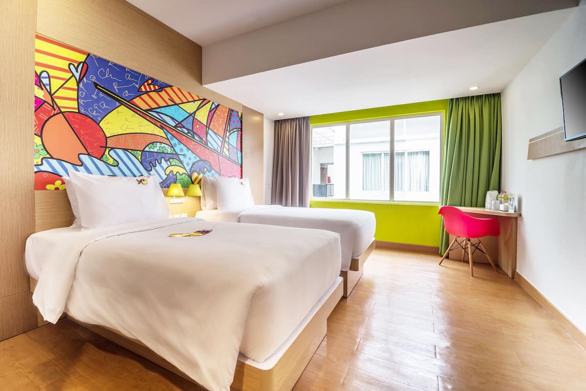 Ein Hotelzimmer mit zwei Betten, einem farbenfrohen Wandgemälde, einem großen Fenster mit Vorhängen und einem Schreibtisch mit Stuhl.