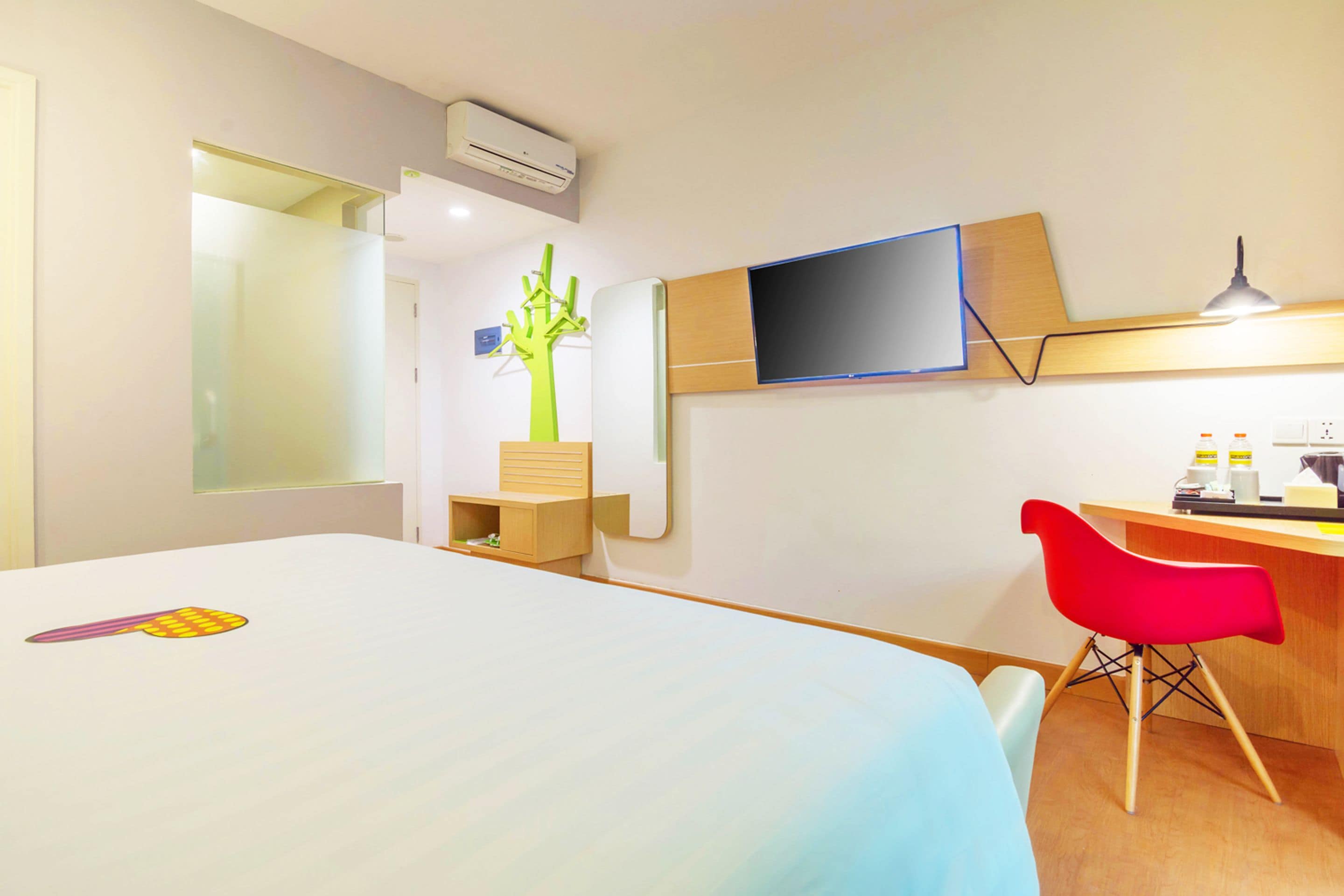 Ein Hotelzimmer mit Bett, Schreibtisch, rotem Sessel, Fernseher und Pflanze.