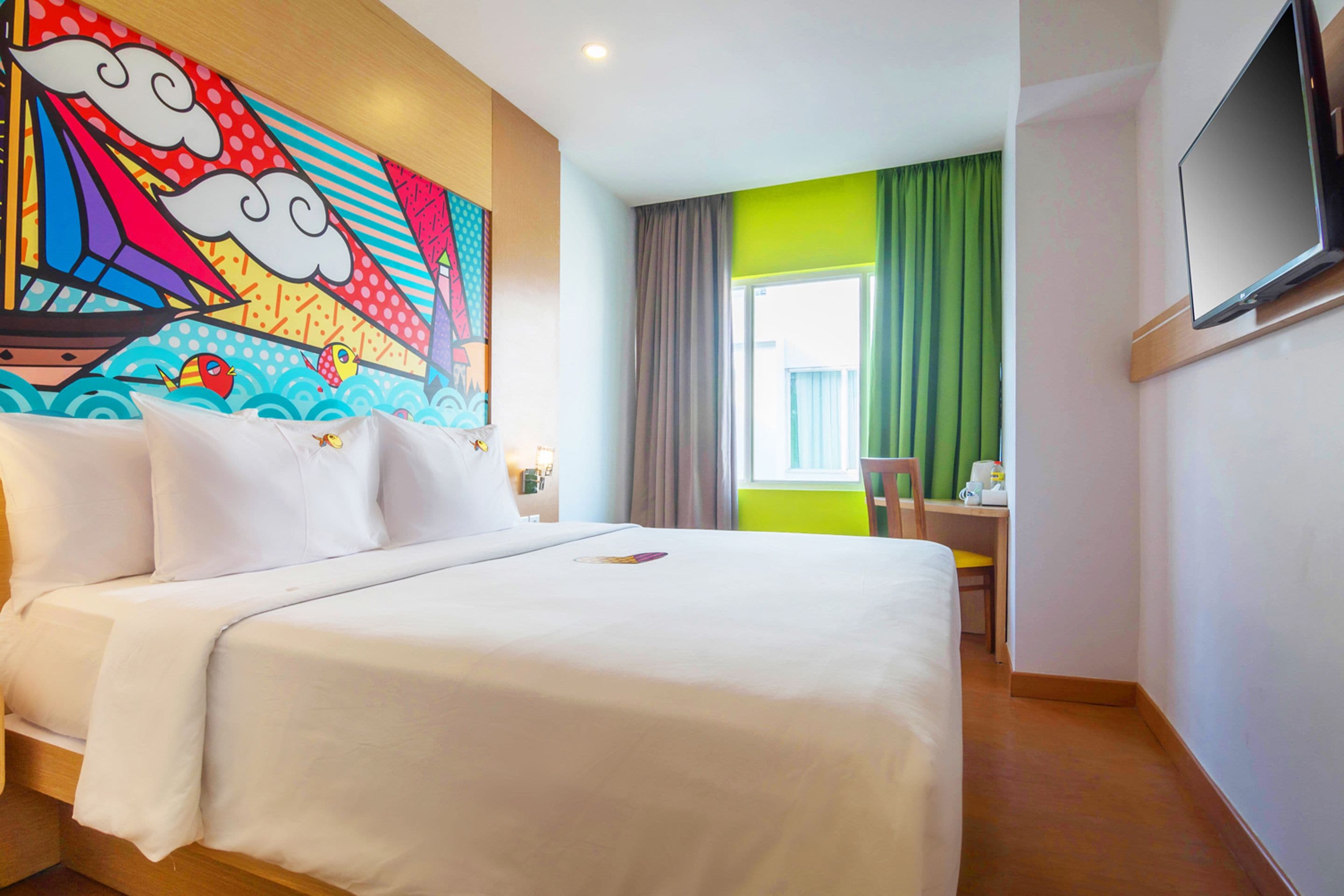Quarto de hotel com uma cama grande, uma escrivaninha, uma cadeira, uma televisão e um mural colorido na parede.