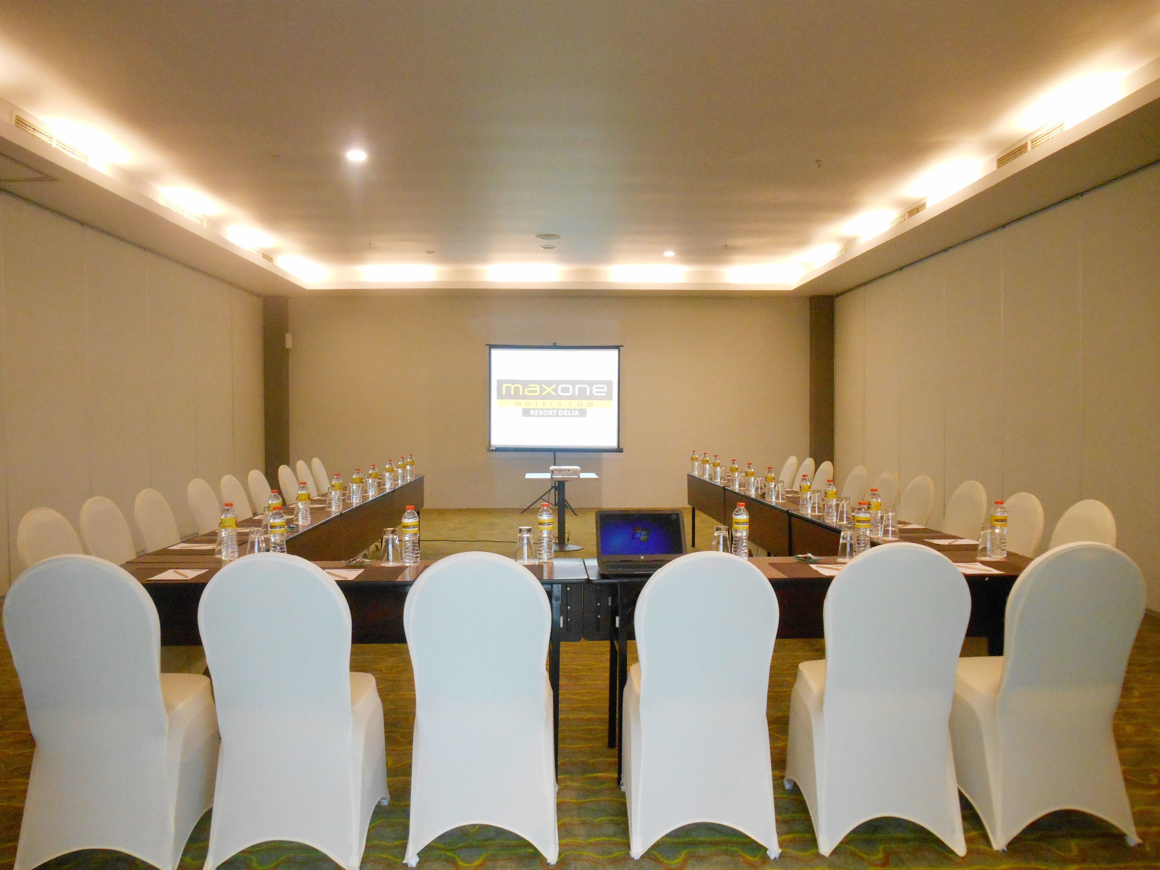 A conference room with a long table, chairs, a laptop, and a projector screen displaying "maxone Hotels.com RESORT DELA".