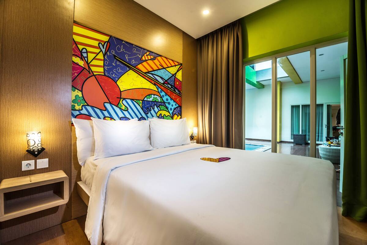 Ein Hotelzimmer mit einem großen Bett, einem farbenfrohen abstrakten Gemälde an der Wand, einem Nachttisch und Blick durch große Fenster nach draußen.