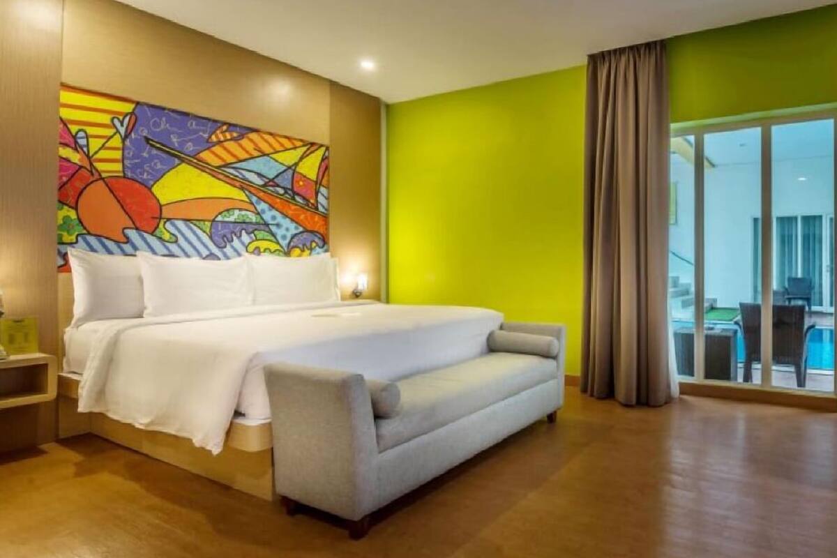 Ein modernes Hotelzimmer mit einem großen Bett, einem grauen Sofa, einem farbenfrohen abstrakten Gemälde an der Wand und Blick auf den Pool durch die Glastür.