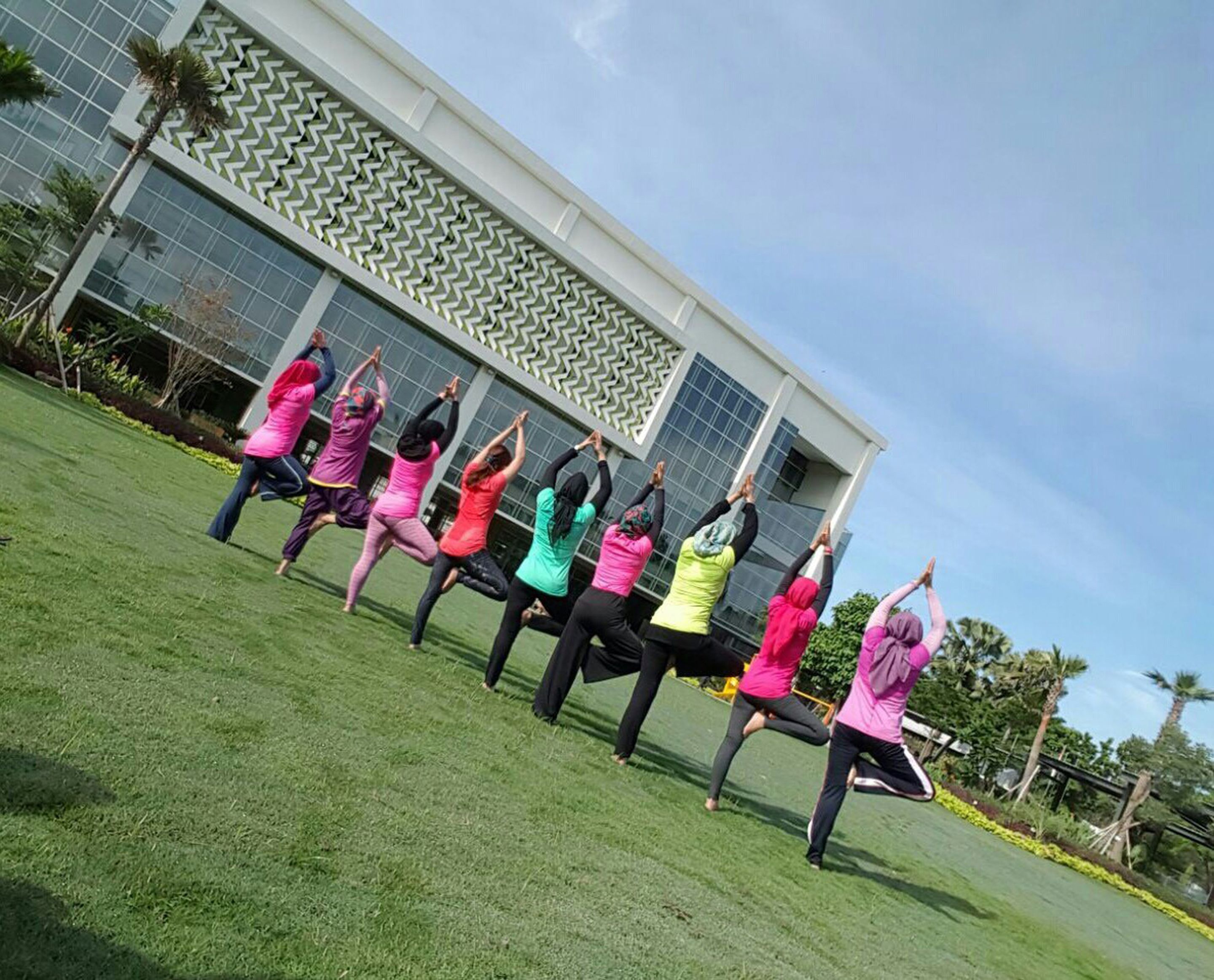 Um grupo de pessoas realiza uma rotina de exercícios sincronizados ao ar livre, em frente a um edifício moderno com fachada estampada.