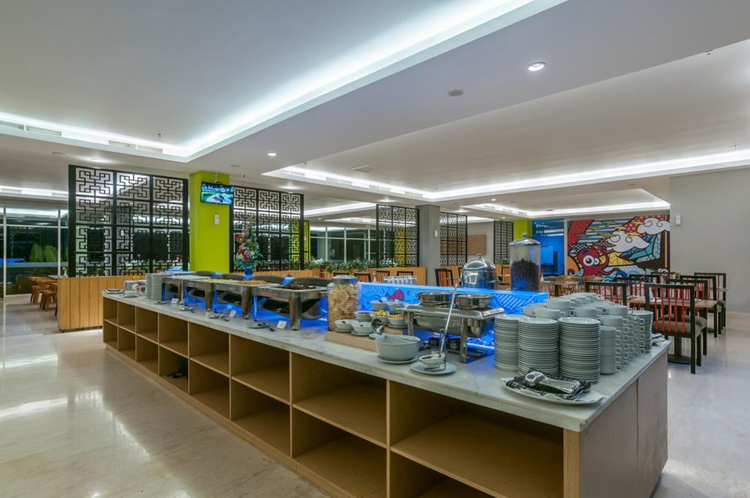 Zona de buffet amb diverses opcions de menjar i estris, zona de seients amb cadires, i elements decoratius com un mural colorit i panells tradicionals.