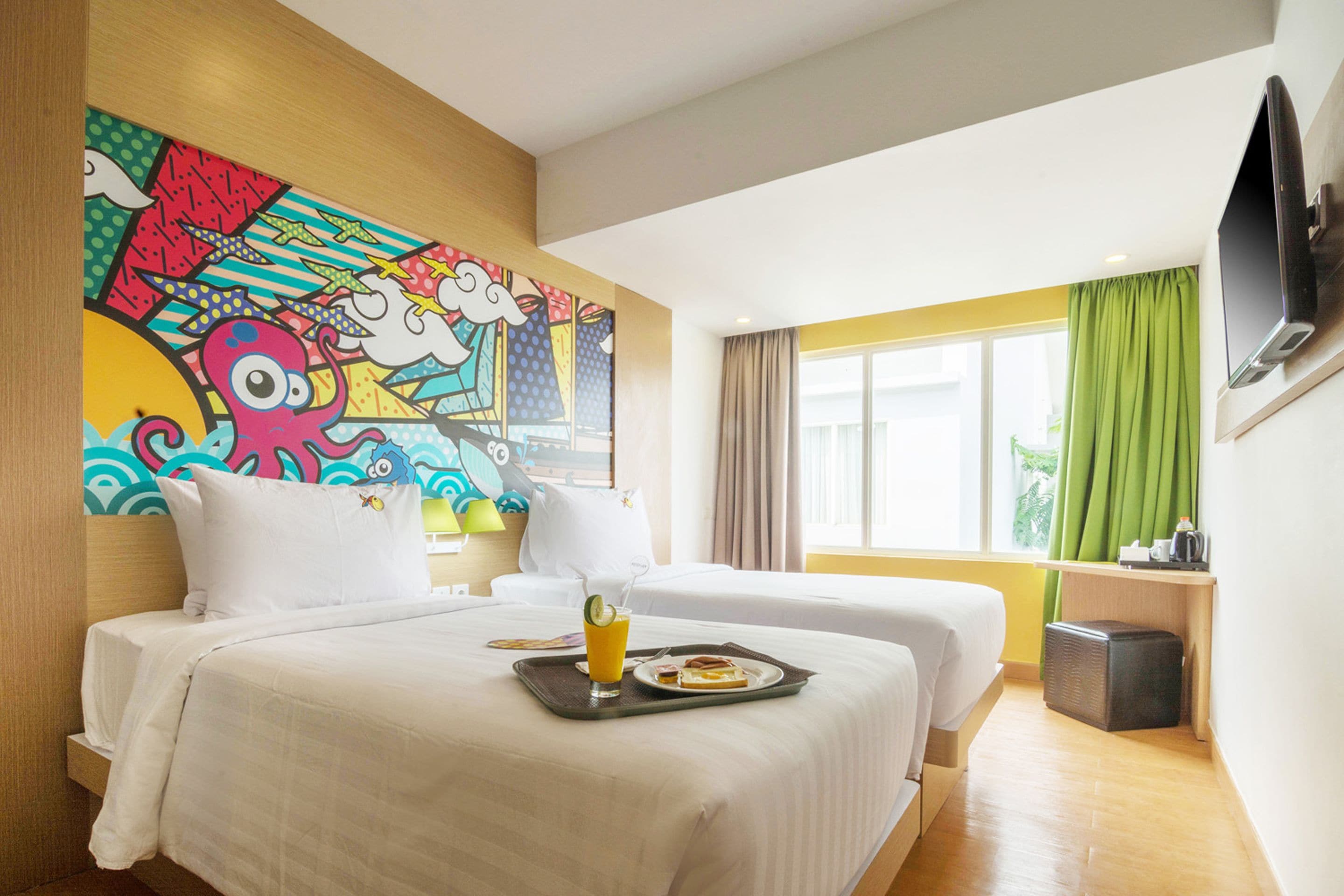 A hotel room with two beds, a colorful mural on the wall, a TV mounted on the wall, and a window with green curtains.