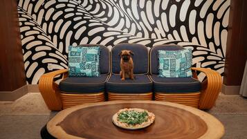 Un perro sentado en un sofá con cojines estampados, una mesa redonda de madera con una pizza al frente, y un diseño abstracto grande en la pared de fondo.