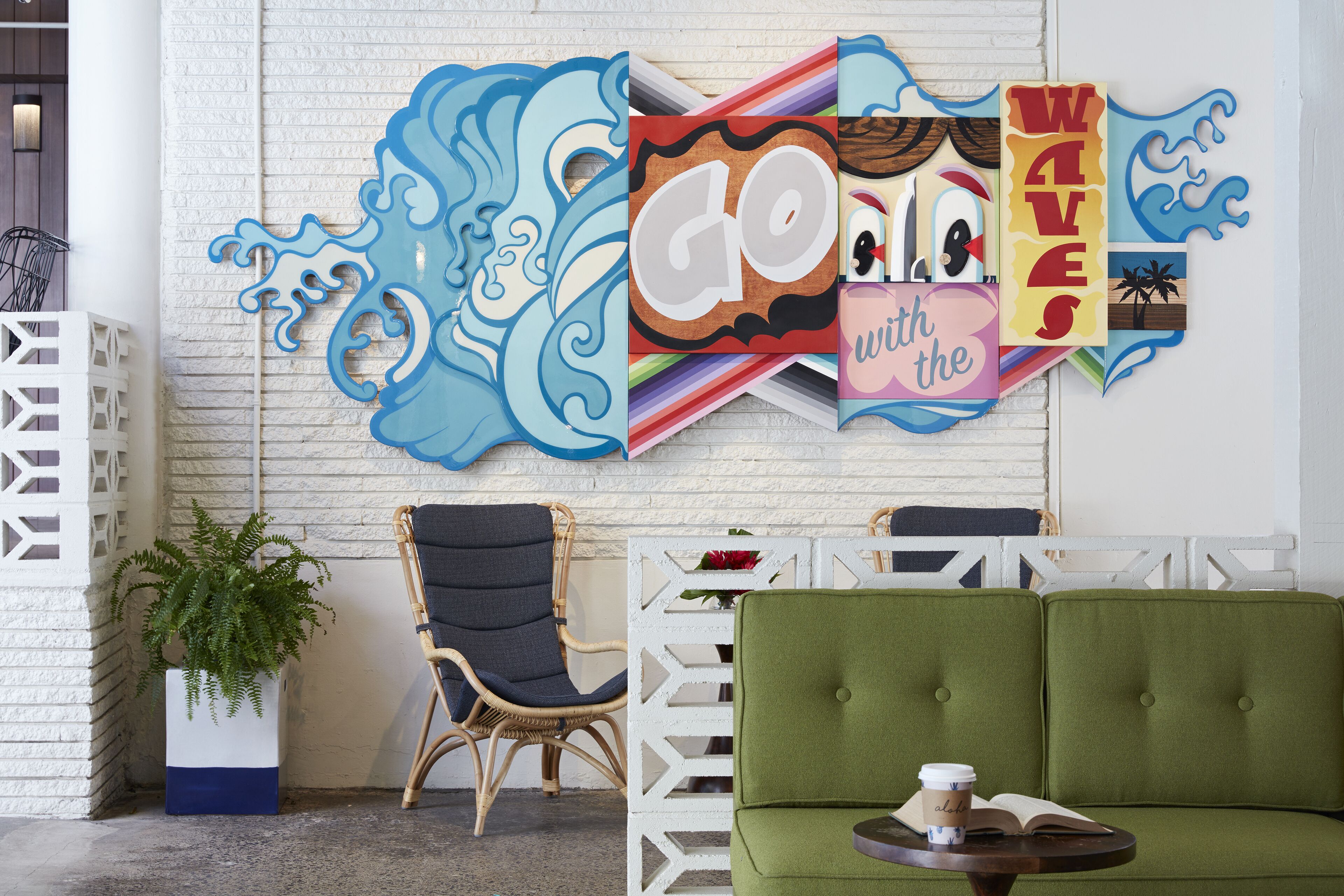 A room with a green sofa, a wicker chair, a white wall with a wave-themed artwork, and a potted plant.