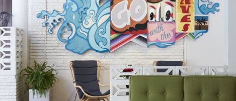 A room with a green sofa, a wicker chair, a white wall with a wave-themed artwork, and a potted plant.