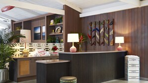 A reception area with a wooden reception desk, two wall-mounted lamps, a small stool, and a set of drawers labeled "Suntrack." - The Surfjack Hotel & Swim Club (Honolulu)