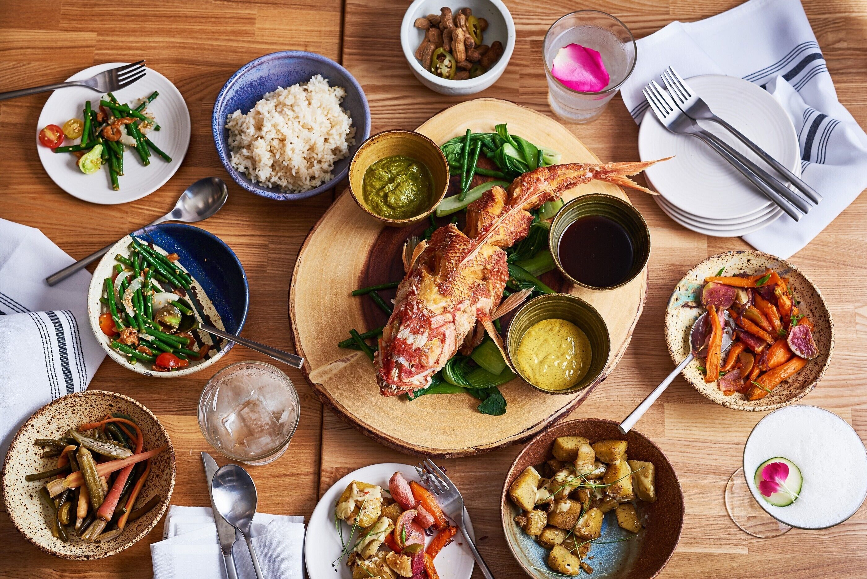 A wooden table with various dishes including rice, vegetables, and a roasted fish.