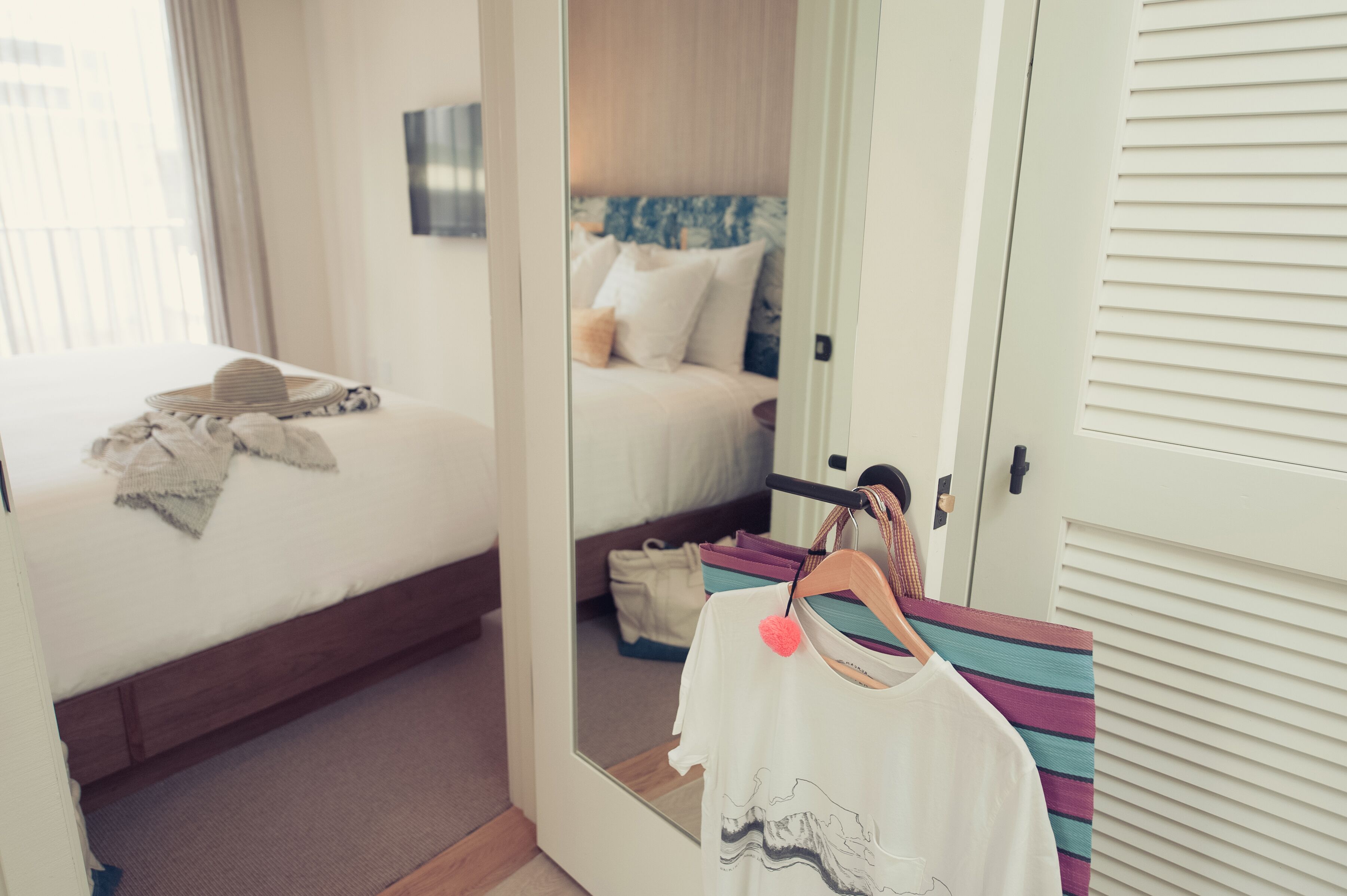 Camera d'albergo con un letto, uno specchio che riflette l'ambiente, un armadio e un appendiabiti con abiti.