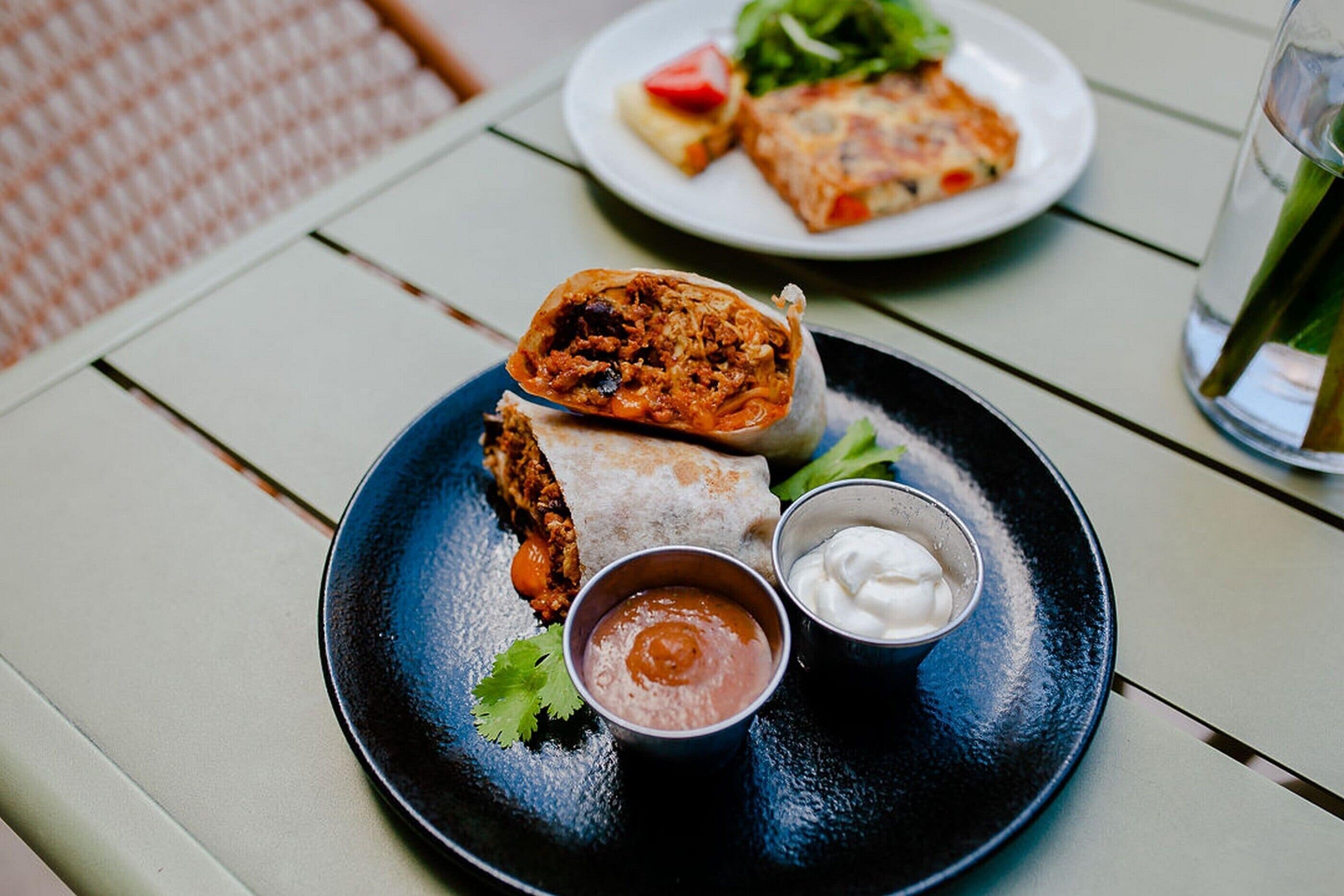 A plate with a burrito, a small bowl of sauce, and a small bowl of sour cream on a table.
