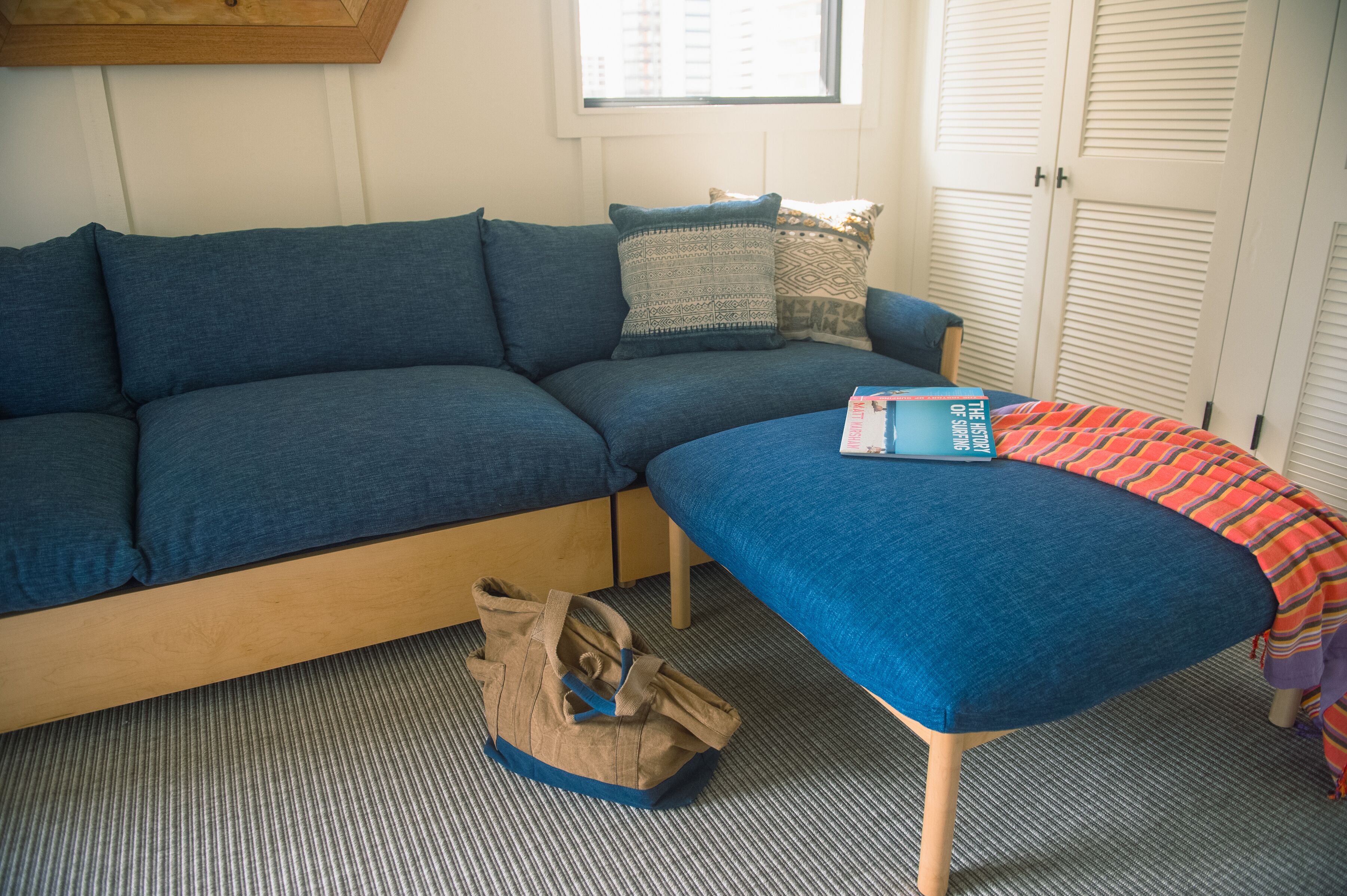 Una habitación con un sofá azul, un reposapiés a juego, una almohada estampada, una ventana y una puerta con contraventanas.