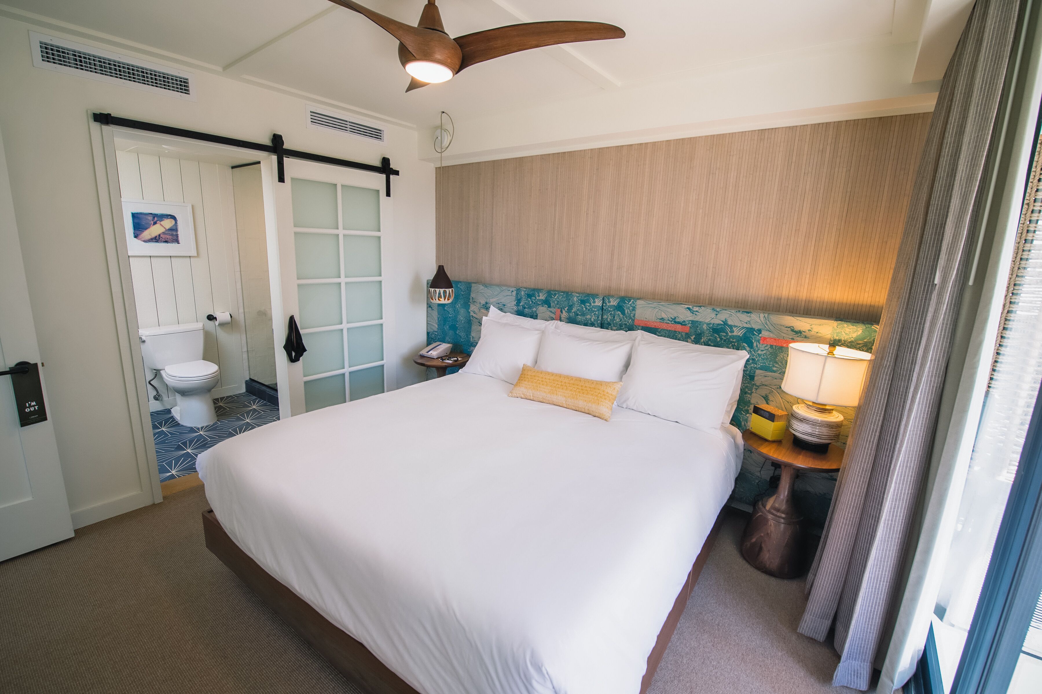 Une chambre d’hôtel avec un grand lit, un ventilateur de plafond en bois, une salle de bain avec des toilettes et un lavabo, et une fenêtre avec des rideaux.