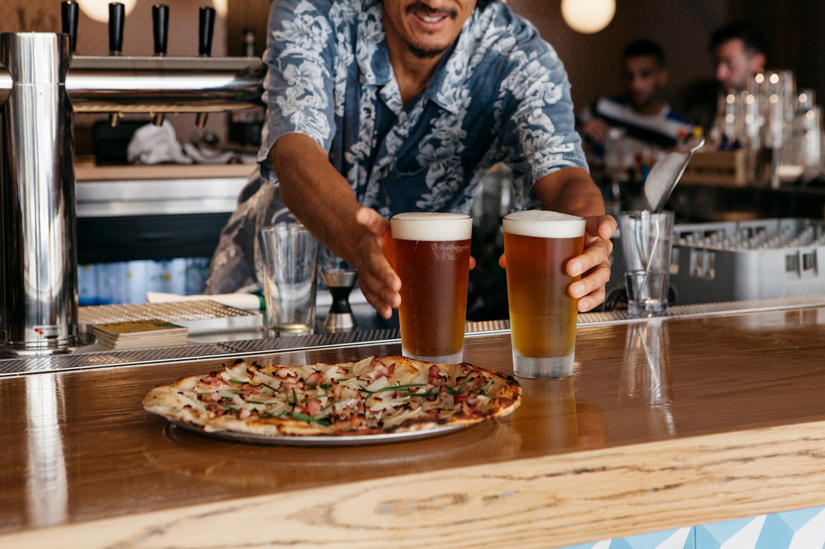 A bar setting with a pizza on the counter and two glasses of beer.