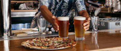 A bar setting with a pizza on the counter and two glasses of beer.