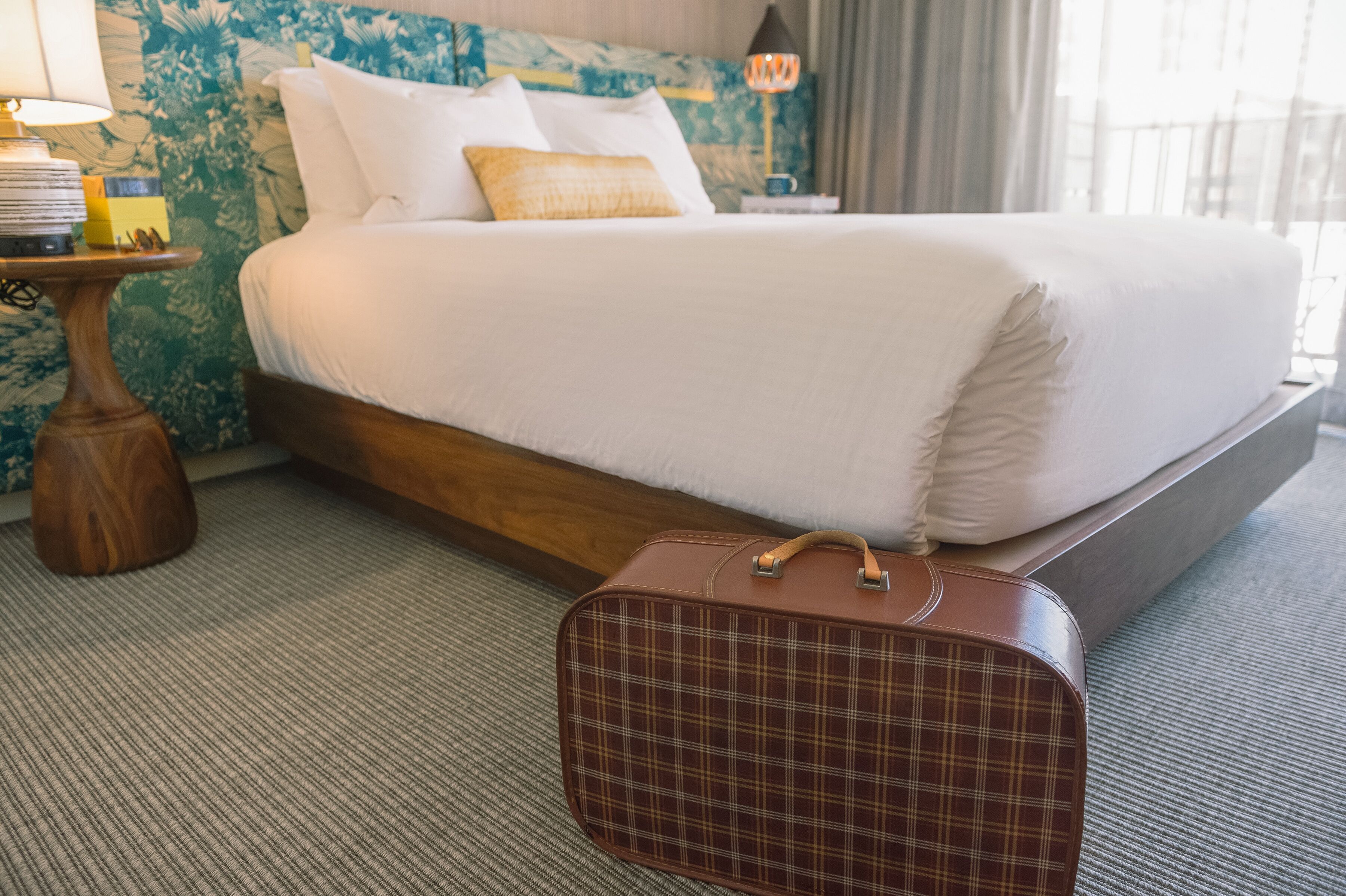 Une chambre d’hôtel avec un grand lit, une table de chevet en bois avec une lampe et une valise à carreaux posée sur le sol.