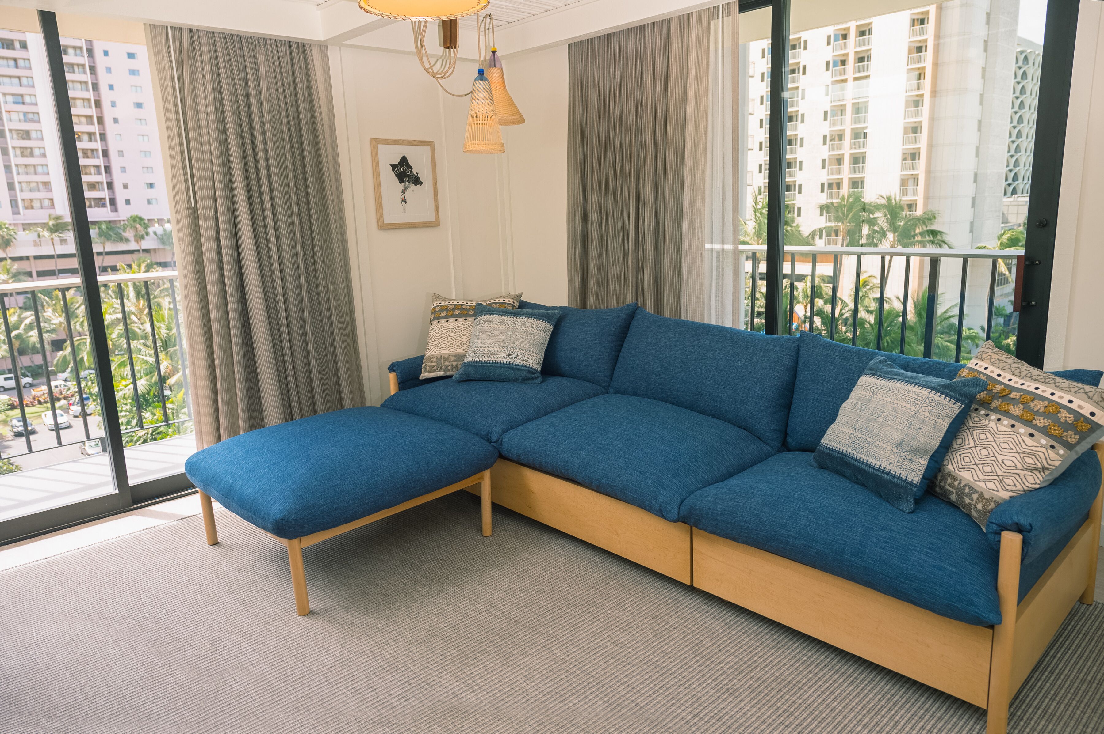 A modern living room with a blue sectional sofa, a small wooden bench, and a large window with a view of a cityscape and palm trees.