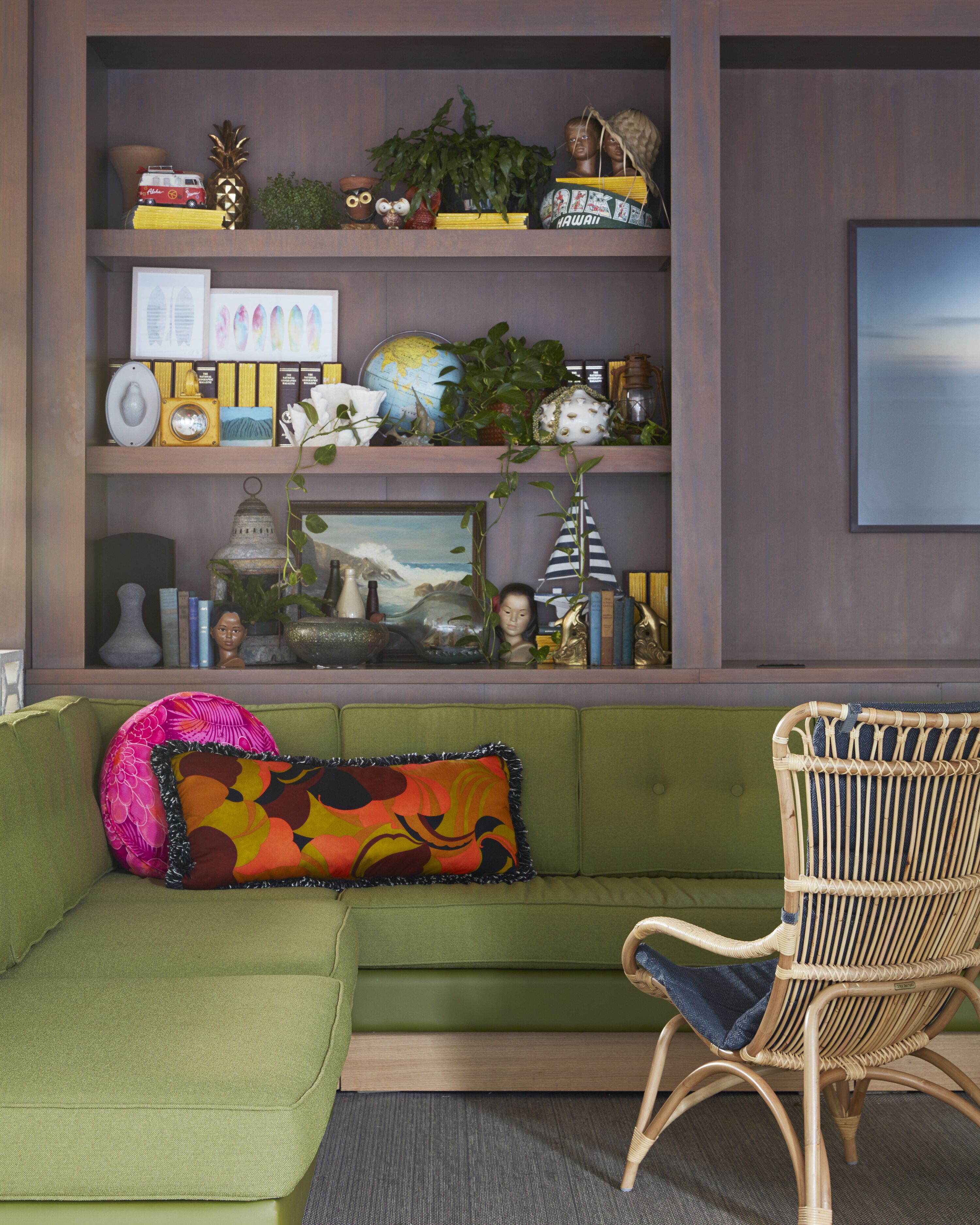 A green sofa with colorful pillows, a wicker chair, and a wooden shelf with various decorative items and a framed picture.