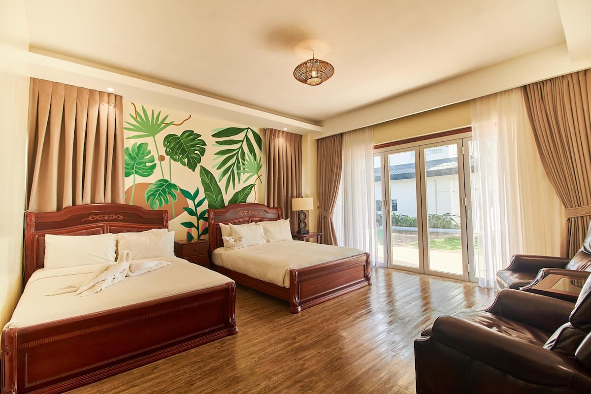 Habitación de hotel con dos camas de madera, un sofá, un ventanal con cortinas y un mural con motivos tropicales.