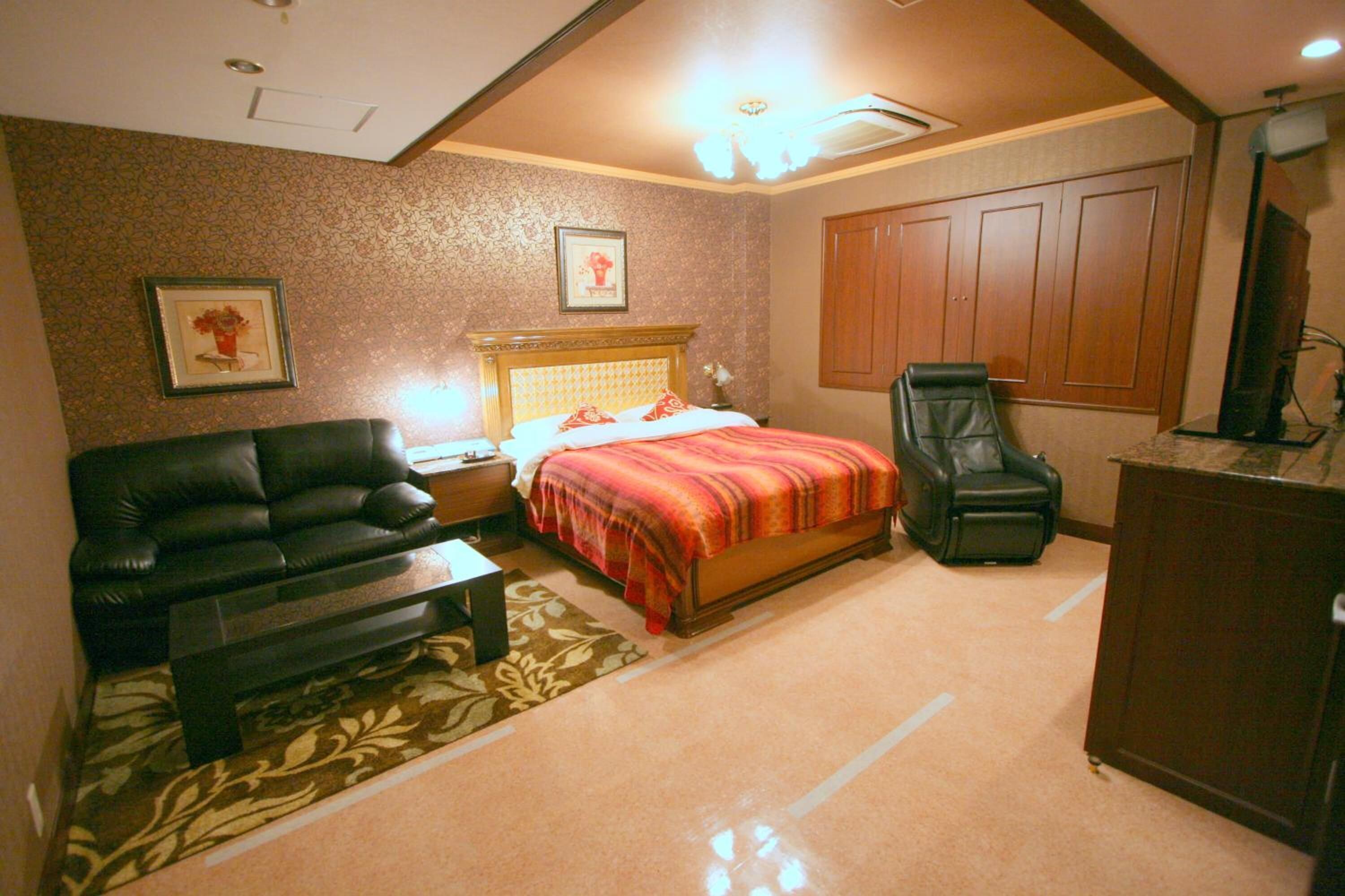 A bedroom with a bed, a black leather sofa, a wooden nightstand, a desk, a chair, and a wardrobe.