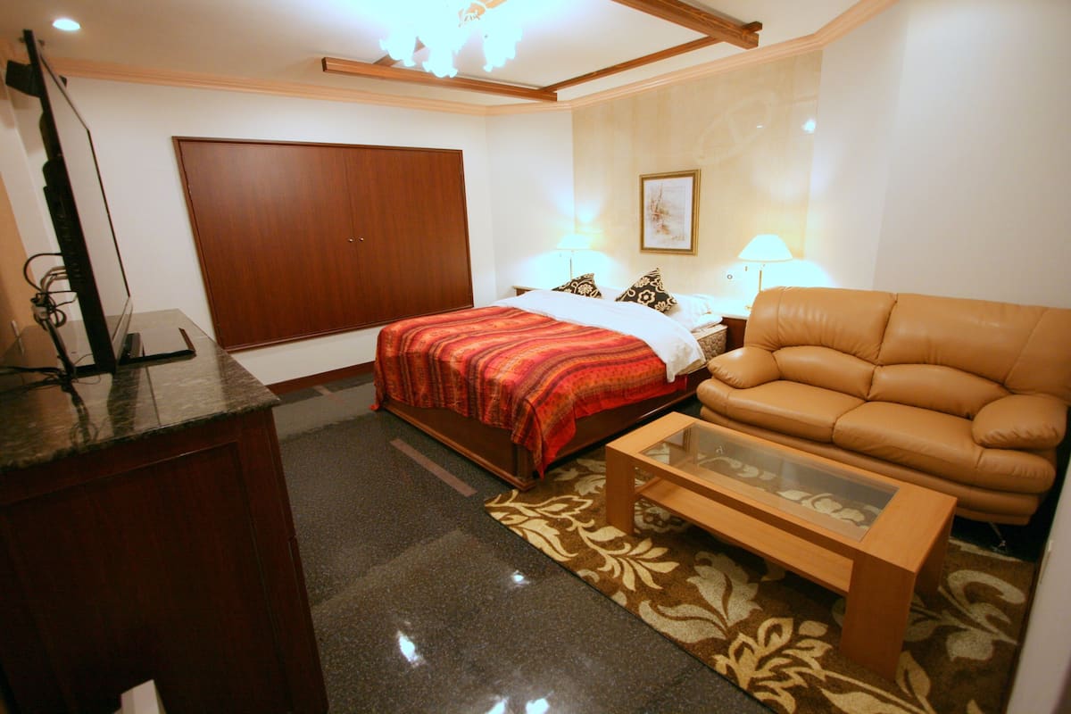 A hotel room with a bed, a leather sofa, a glass-top coffee table, a desk with a computer, and a large wardrobe.