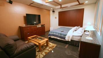 A compact hotel room with a sofa, a television, a bed with a patterned bedspread, a bedside table, and a lamp.