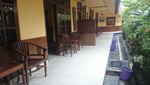 A covered outdoor seating area with wooden benches and tables, a tiled floor, and a planter with plants and purple baskets. - Handika Hotel Mataram (Mataram)