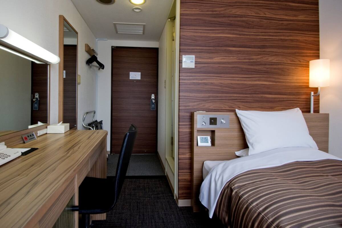 Une chambre d’hôtel avec une tête de lit en bois, un lit avec une literie à rayures blanches et marron, un bureau avec un miroir, une chaise et une lampe.