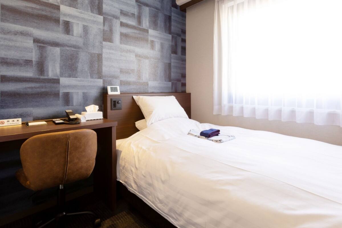 Une chambre d’hôtel avec un lit, un bureau, une chaise, une table de chevet et une fenêtre avec des rideaux.