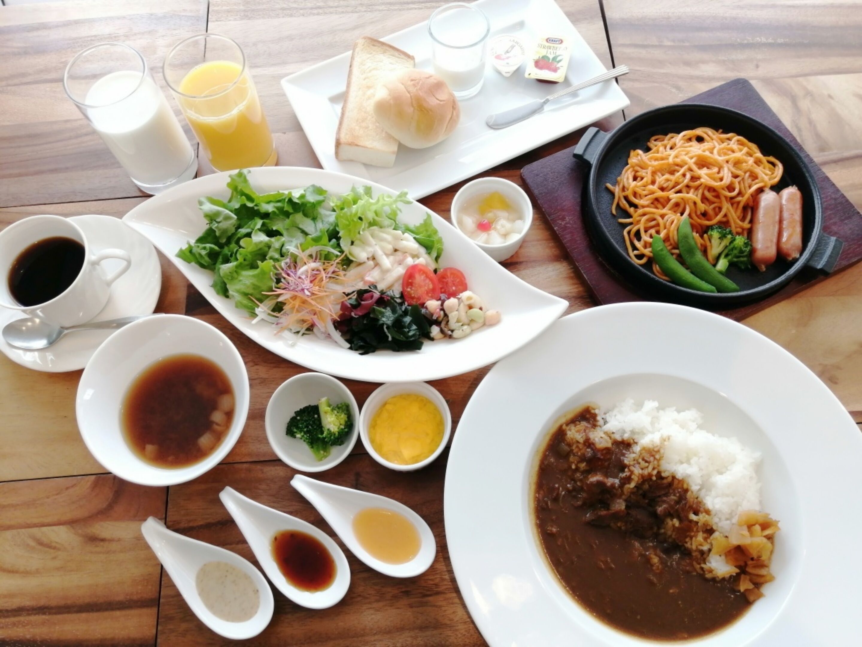 A table with various dishes including a salad, noodles, rice, and soup. There are also glasses of milk and juice, and a plate with bread.